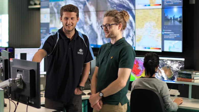 Two young men in front of computer screen with large screens of National Operations Centre behind them.