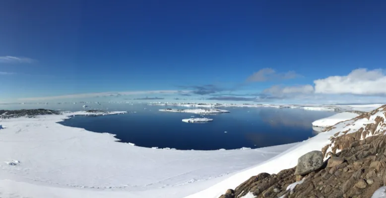 Summer landscape with ice and snow in foreground, looking out onto blue water under a mostly clear blue sky.