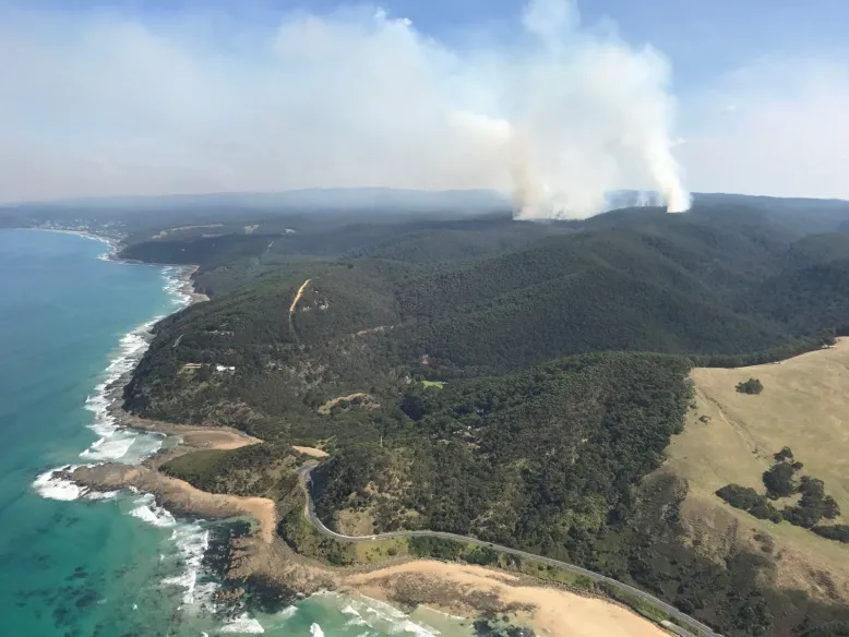 Aerial photo looking from the ocean over the coastline to forested hills in Victoria's Otway region. Smoke rises from a prescribed burn at Big Hill Track.