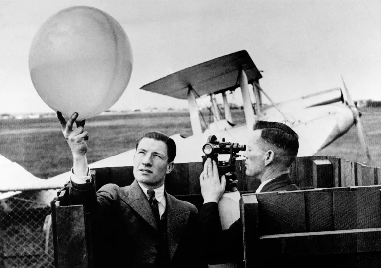 Man holds up weather balloon while another man looks through a theodolite towards it. In the background is a Gypsy Moth plane.