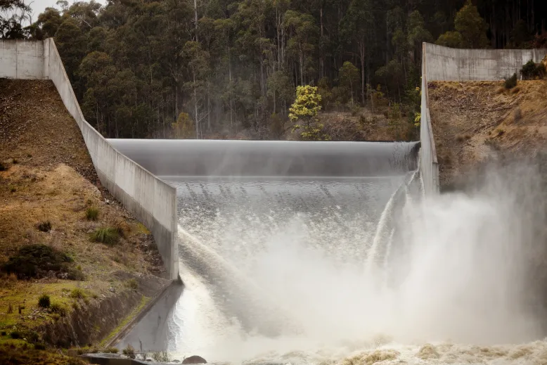 Photo of water surging over a dam edge into a narrowing v-shaped spillway. Spray rises as it hits the bottom. On the far bank of the dam, bushland rises up a steep incline.