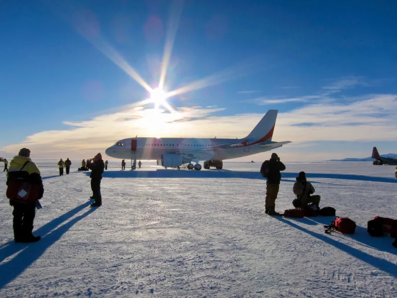 Passengers prepare to board a small plane on the icy Wilkins Runway, under a bright blue sky.