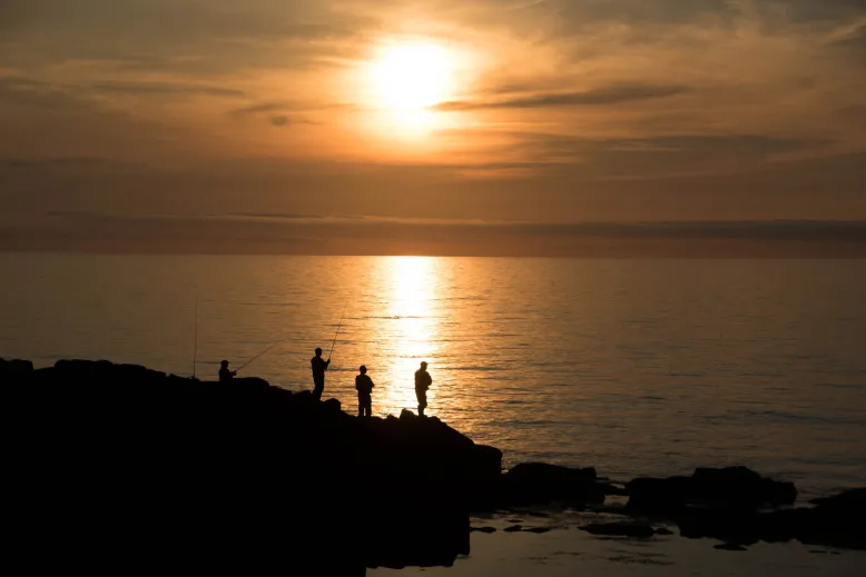 Group of people fishing from a rock ledge on the coast. Sun is setting in the background, casting soft orange light across the calm ocean.