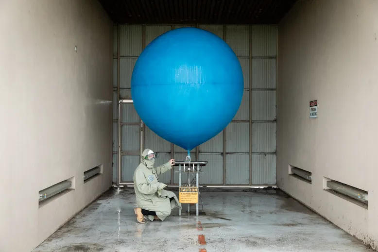 Person in protective equipment kneeling while filling a weather balloon with hydrogen gas. The balloon is blue and bigger than the person.