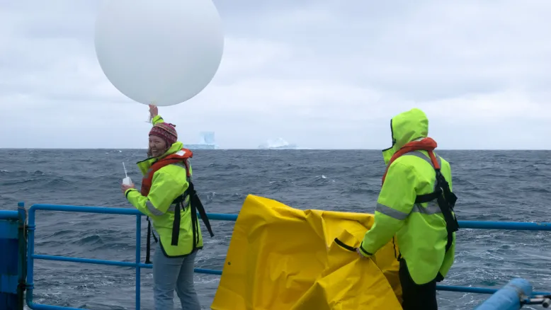 Two scientists in hi-vis vests release a weather balloon into the Southern Ocean atmosphere from the deck of a ship. On the left, a female scientist on the left is holding the balloon and a communications device. Another scientist on the right is holding a yellow tarpaulin.