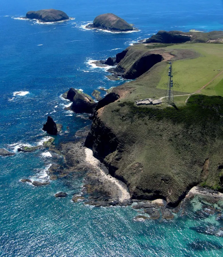 Aerial photo of Kennaook / Cape Grim station on the tip of a green but rocky cliff, above a turquoise and blue ocean.