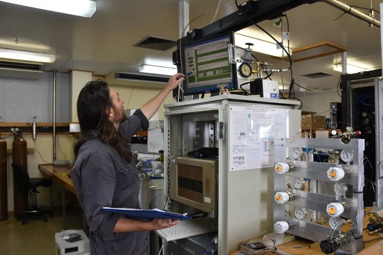 Man reaches up to computer screen showing data. It's on top of a grey box containing another screen. There is more equipment with handles and dials to the right.
