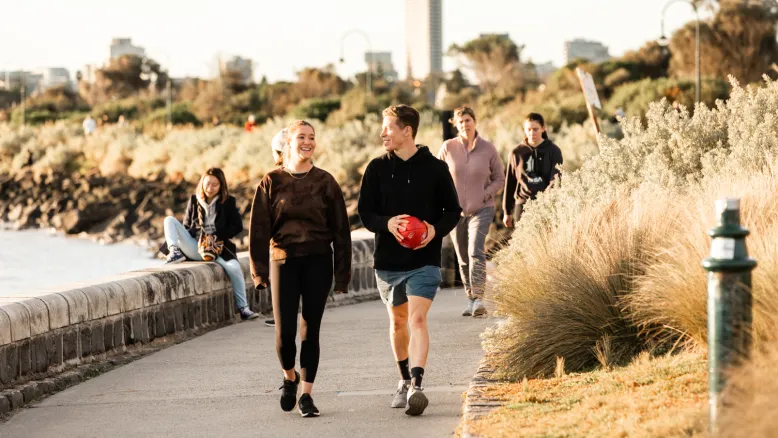 Two young people walking in the late afternoon sun along Elwood beach, smiling. One is holding a footy.