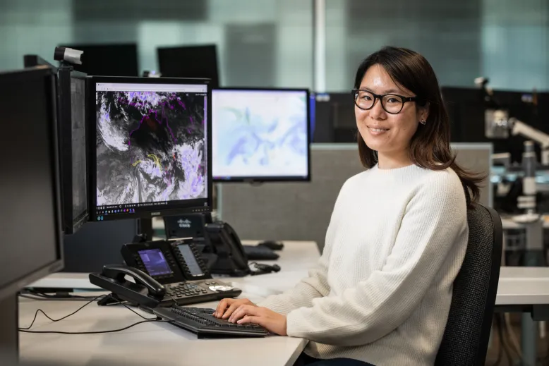 Woman sitting at a desk in an office with her hands on a computer keyboard. She is smiling. The computer screens show weather systems swirling around a satellite image of Australia.