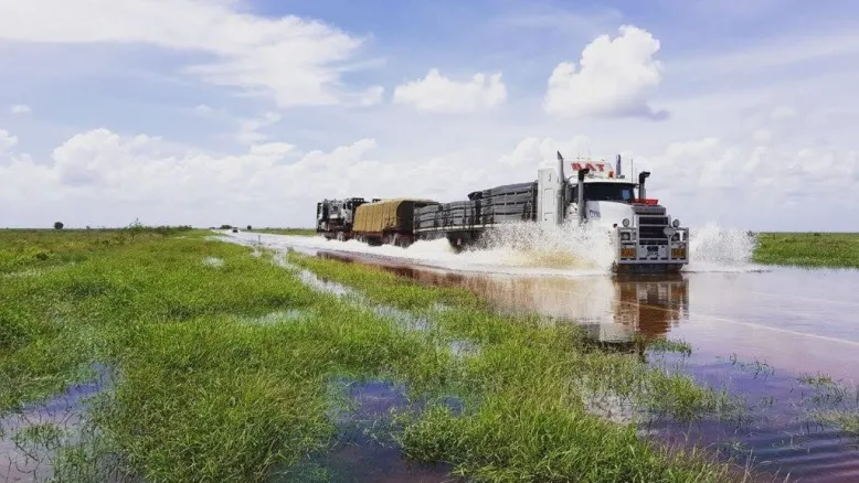 Heavily-laden road train drives through floodwater across a road under a pale blue bright sky.