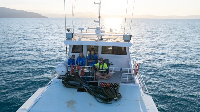 Four people sitting on a boat, sailing a calm sea under blue skies. There is an island in the background.