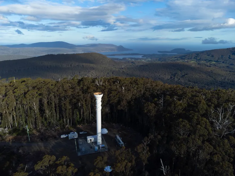 White radar tower without a radome on top of a heavily forested mountain.