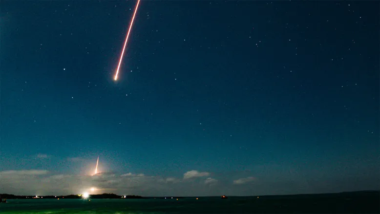 Bright rocket trail streaks upwards through a starry evening sky above the launch field.