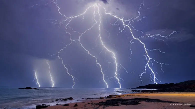Huge, sprawling lightning strike covers the sky over a rocky beach.
