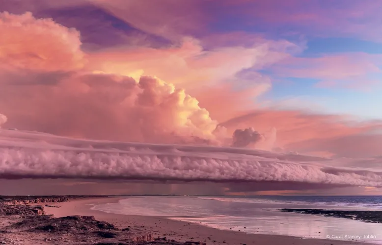 Long, flowing clouds lit by the twilight sun roll across the sky over a rocky beach and out onto the ocean.
