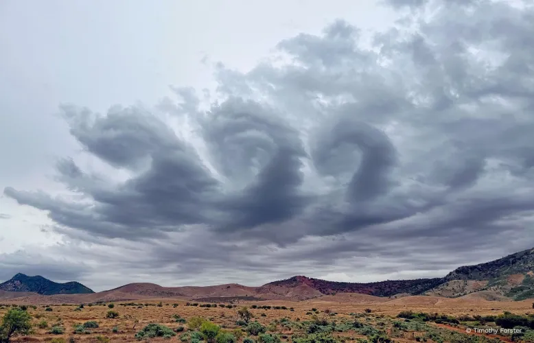 Clouds whirl like waves crashing on the shore above mountainous terrain.