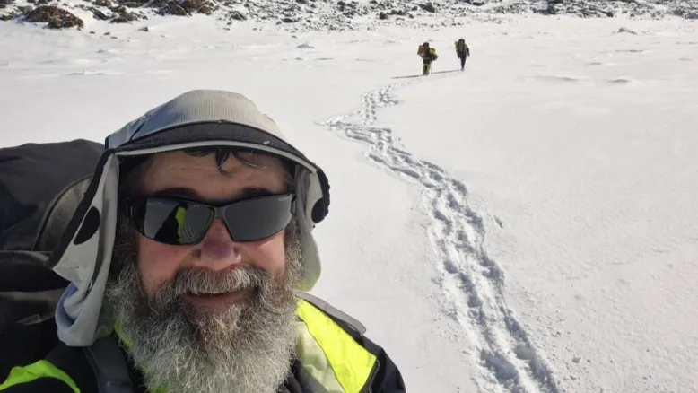 Technical officer Brian takes a selfie on the snow with two colleagues walking in the distance behind him