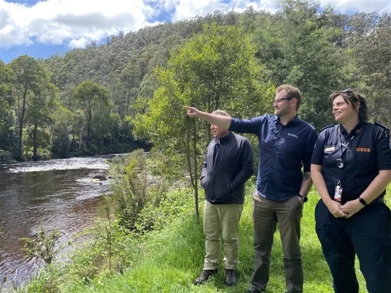 Meteorologist in middle pointing across a river with one SES staff on each side of him.
