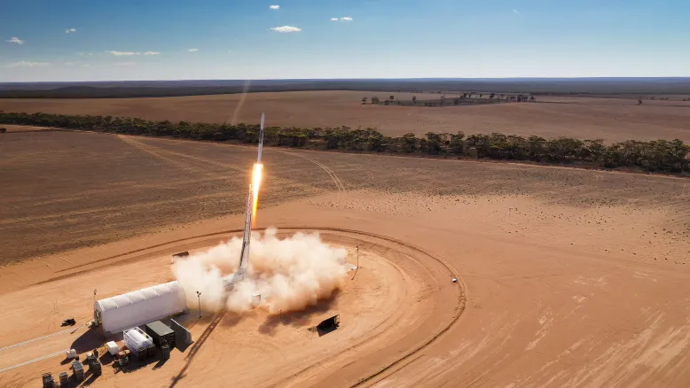 Rocket lifts off from a launch pad in an arid landscape, leaving a cloud of dust as it rises into a blue sky.