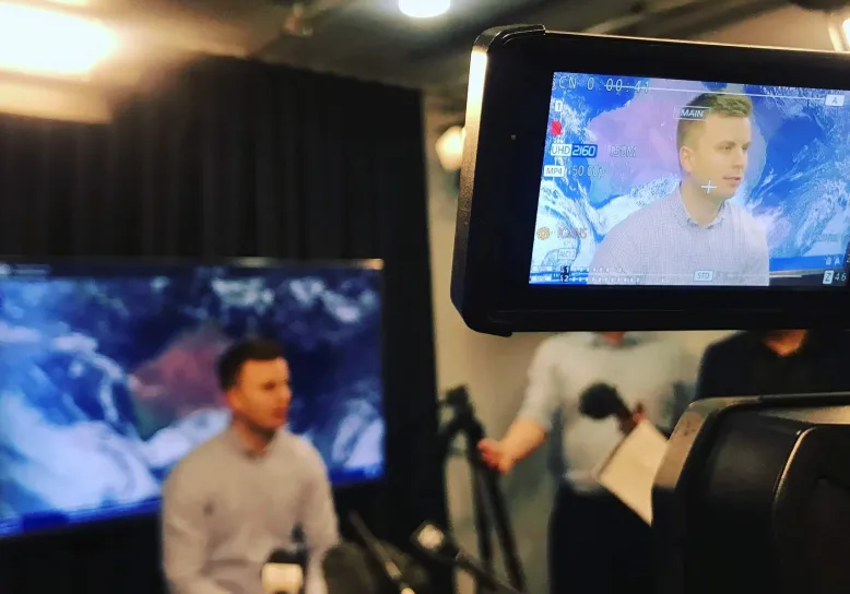 Meteorologist Harry Clark sitting in front of a screen showing an image of Australia with a camera filming for a television interview.