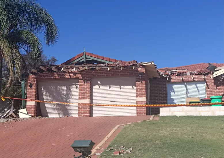Red brick suburban house with a severely damaged, partially collapsed roof, and debris scattered around the driveway. Caution tape is stretched across the front under a clear blue sky.