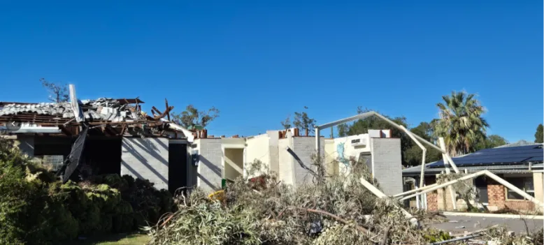 White brick house with tornado damage including a destroyed roof, bent steel frames and tree branches.