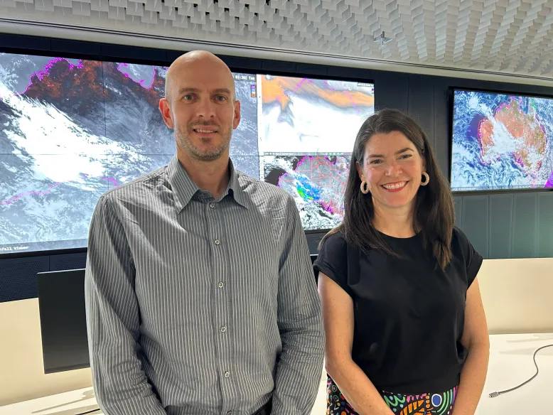 Two bureau researchers standing in a weather operations centre with large screens in the background showing satellite and weather maps.