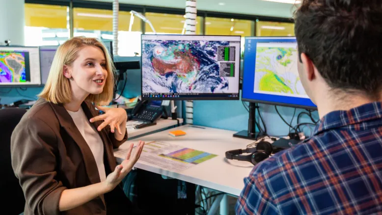 Two people discussing weather data at a workstation with multiple meteorological maps displayed on computer monitors.