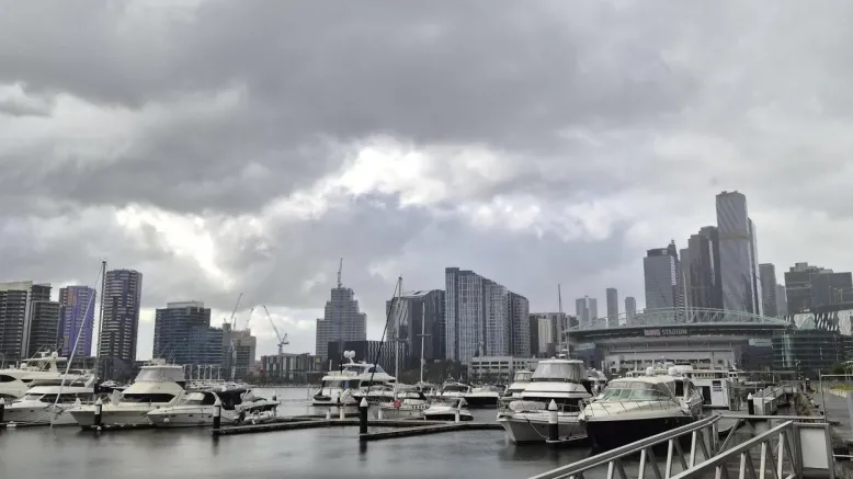 Docklands marina with moored boats and Melbourne city skyline beneath dark, low cloud and patches of brighter sky.