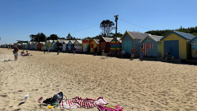 Colourful beach bathing boxes lined up along a sandy beach with people nearby under a clear blue sky.