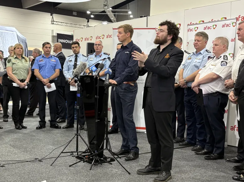 Three people speaking at a media conference. A group of emergency services "Three people speaking at a media conference. A group of emergency services personnel stand behind them.