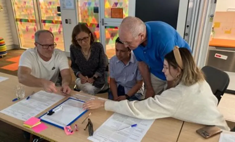 Five people of various ages gathered around a table where they are working collectively on a task on paper.