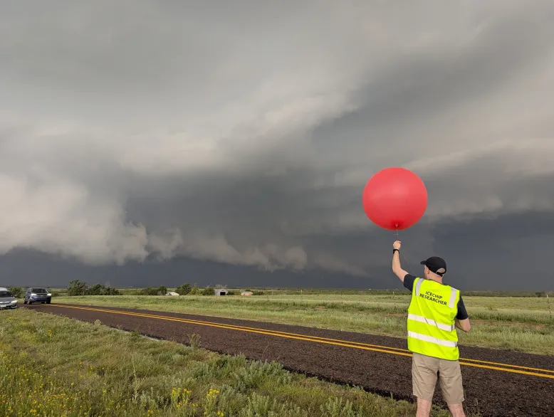 Man in a high vis vest holding a red observation balloon. In the background is a dark thunderstorm.