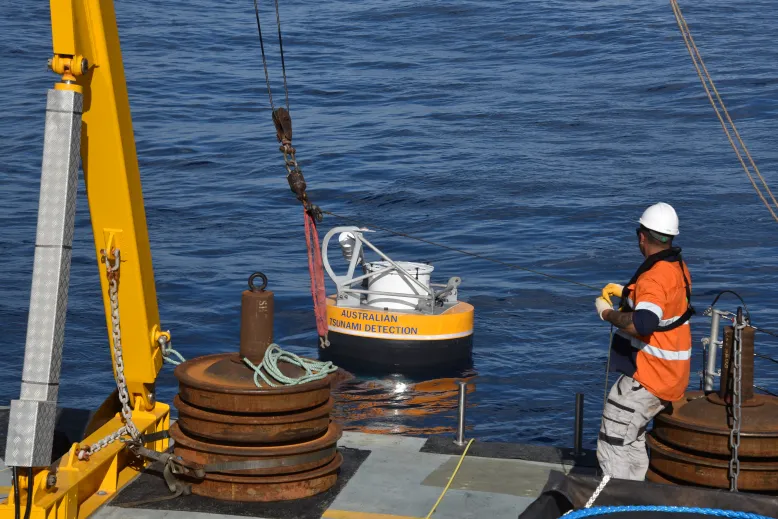 Large tsunami observation buoy being lowered into the ocean from a vessel using a crane and lifting rig. A crew member assists with the deployment.
