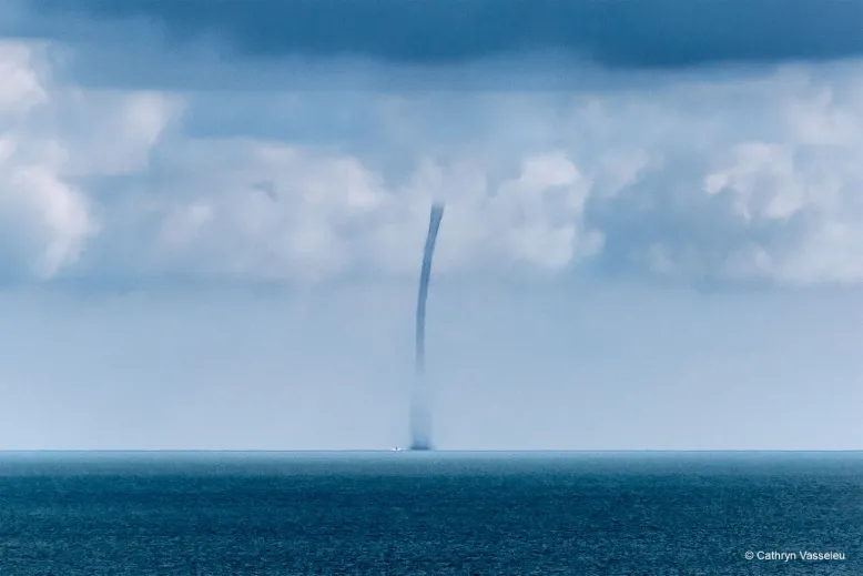 Dark blue waterspout rising from a calm ocean. The waterspout connects to white clouds high in the sky.