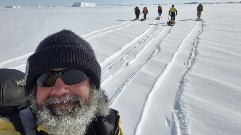 Man wearing a beanie, sunglasses and a frost-covered beard stands in a snowy landscape, with sled tracks leading to a group trekking toward icebergs in the distance.