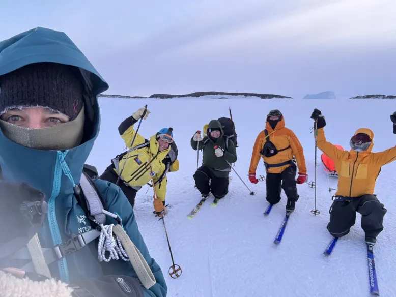 Five people on skis in a snow-covered frozen landscape, with low rocky hills visible in the distance.