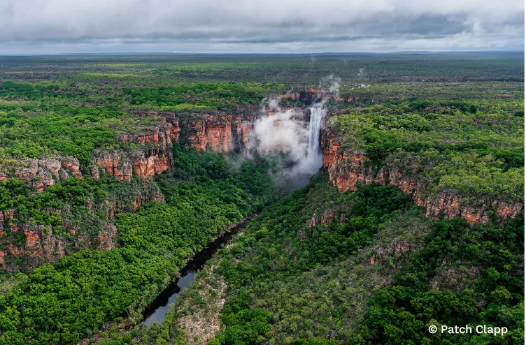 Waterfall plunges into a mist-filled gorge surrounded by dense forest and towering red cliffs.