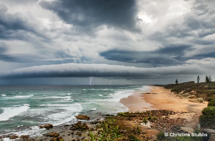 Long, low roll cloud advances over a rugged coastline while lightning strikes from a stormy sky.