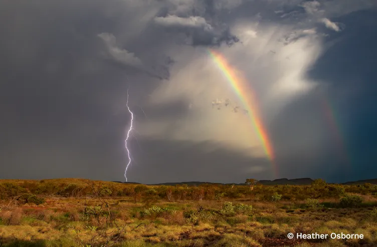 Rainbow glows beside a lightning strike as storm clouds gather over rugged grassland.