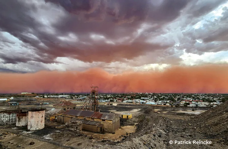 Wall of red dust advancing across a mining town beneath towering dark storm clouds.