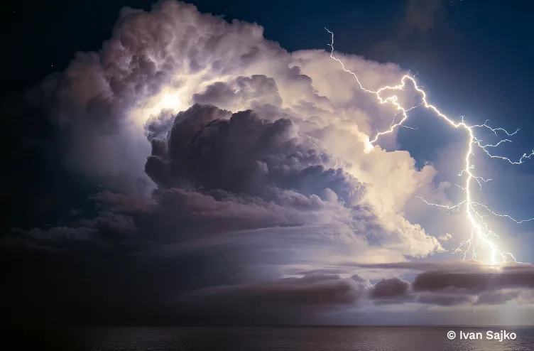 Forked lightning illuminates a towering thunderstorm cloud above a dark and tranquil sea.