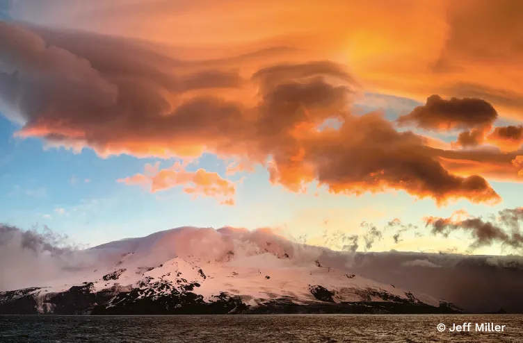 Snow-covered volcanic island sits beneath sweeping orange lenticular clouds that blaze across the sky at sunset.