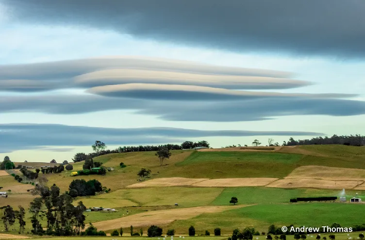 Stacked lenticular clouds hover above a patchwork of rolling green and golden fields beneath a brooding sky.