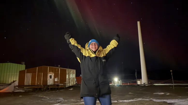 Person stands outdoors with their arms held up in the sky beneath colourful aurora lights in the night sky.