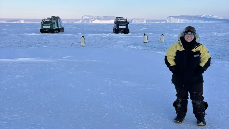 Person in cold-weather gear standing on sea ice. Penguins, tracked vehicles and icebergs are visible in the background.