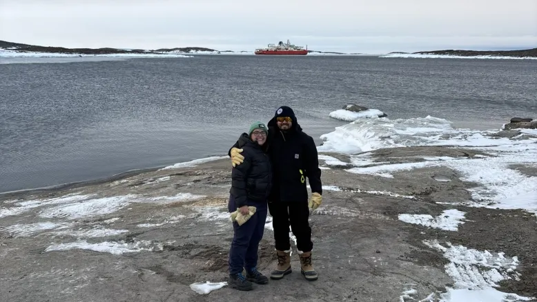 Two people standing on a rocky Antarctic shoreline beside icy waters, with the ship Nuyina visible in the distance.