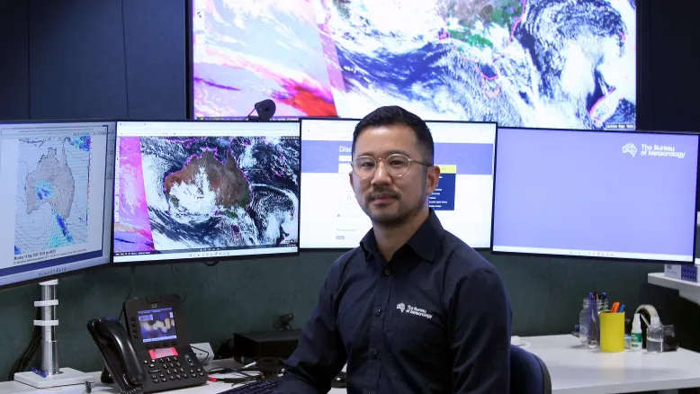Meteorologist at desk in front of screens displaying satellite weather images.