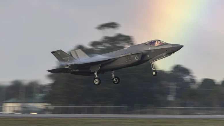 Fighter aircraft with wheels down, about to land on an airstrip. A rainbow colours the sky behind the cockpit.
