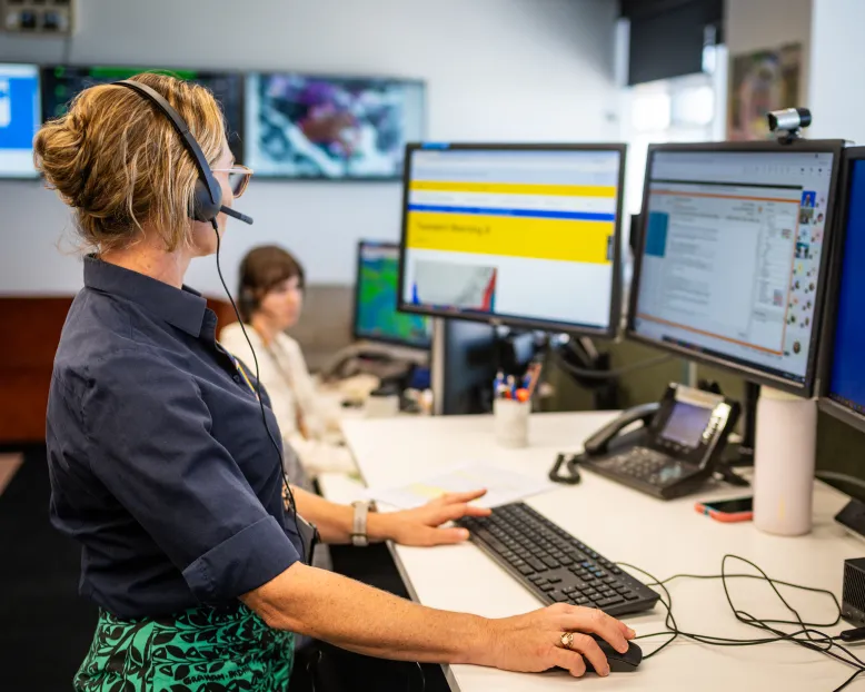 Female meteorologist standing at desk looking at two computer monitors with another woman in the background.